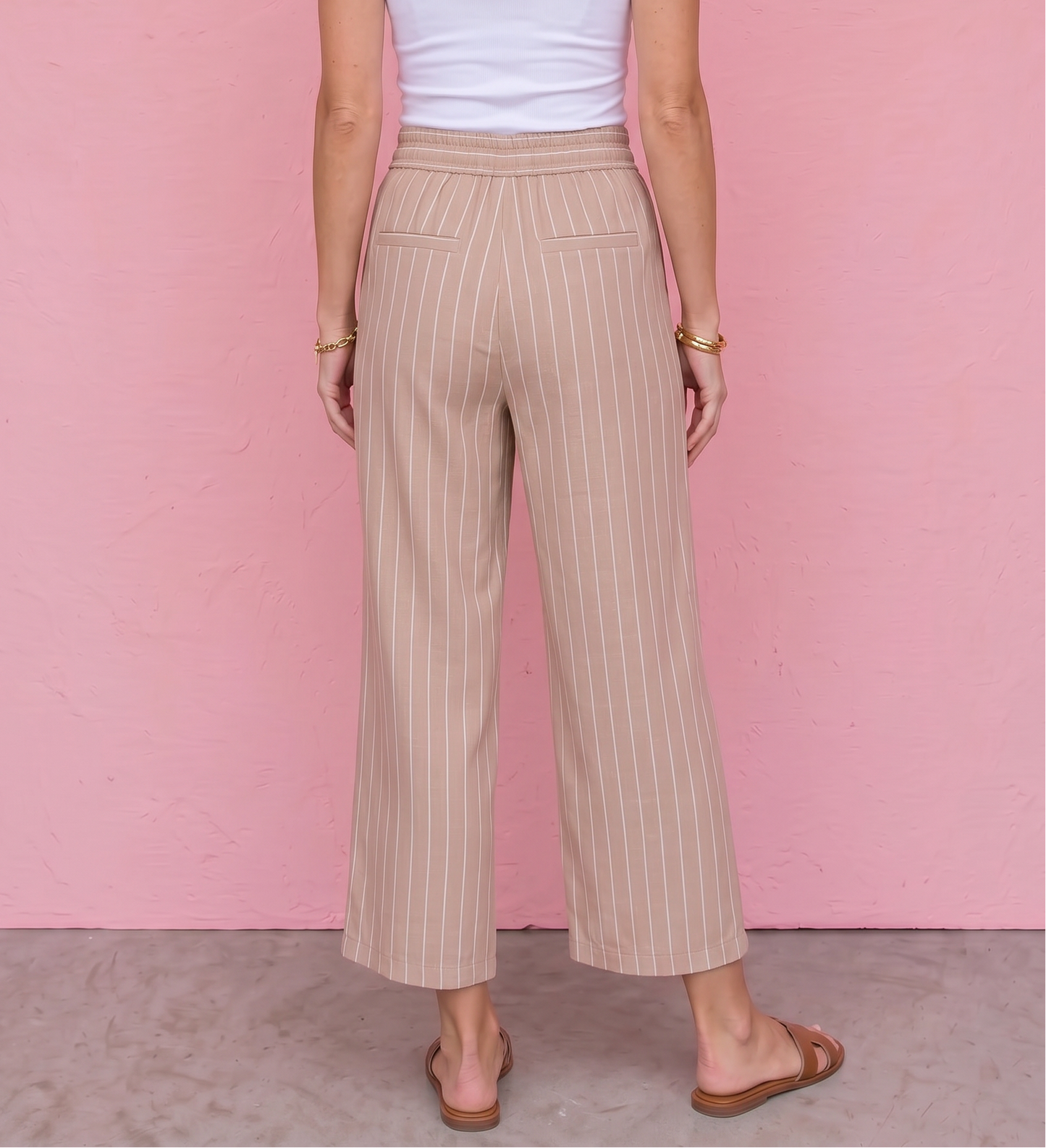 Woman wearing a white tank top and beige striped pants against a pink wall.