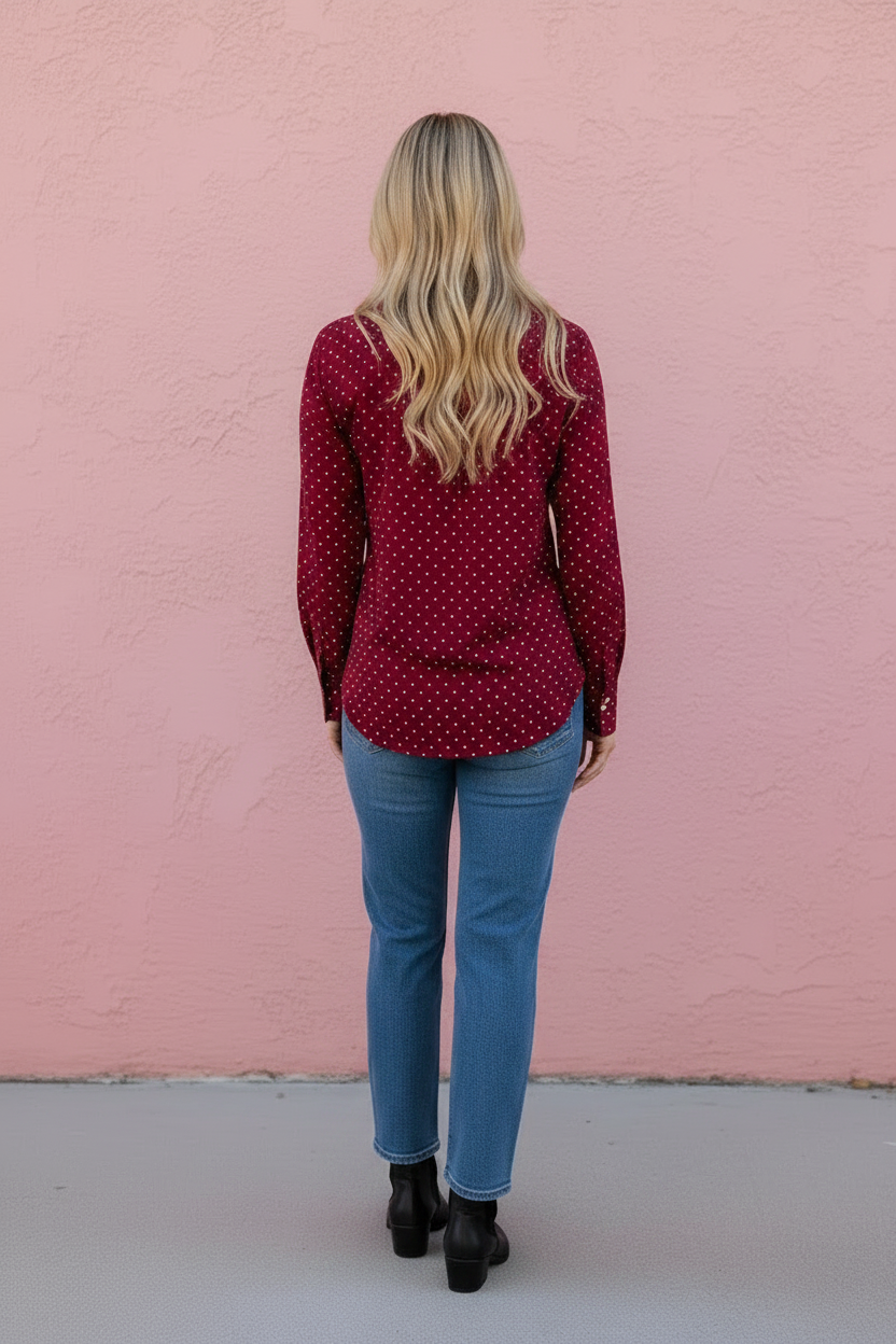 A woman with blonde hair is shown from behind, wearing a dark red long-sleeve button-up shirt with a small white polka dot pattern, blue jeans, and black ankle boots, standing against a pink wall.