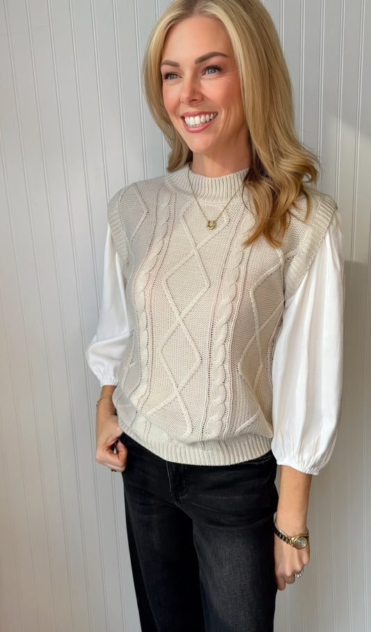 A smiling blonde woman wearing a cream cable-knit sweater vest over a white long-sleeve blouse with dark jeans, standing against a white paneled wall.