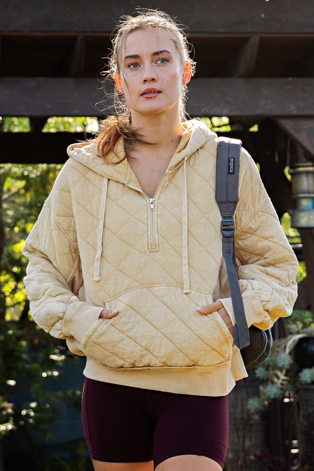 A woman wearing a quilted, half-zip hooded sweatshirt in a light beige or cream color, paired with dark purple athletic shorts and a black backpack, standing outdoors under a wooden structure.