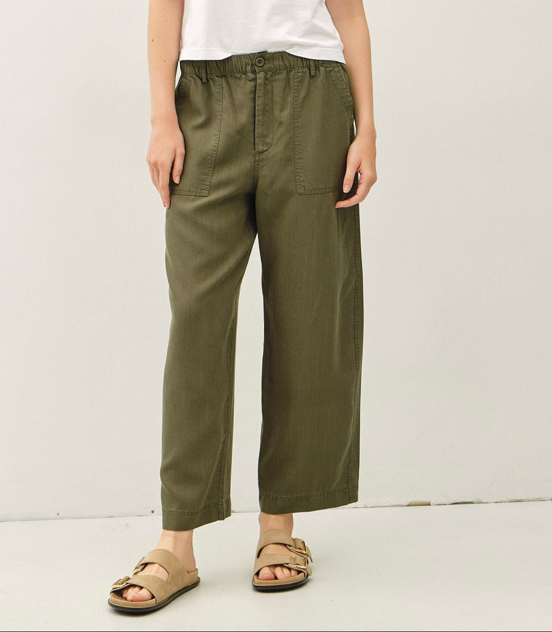 A woman modeling a white short-sleeve crewneck t-shirt and olive green wide-leg cropped pants, paired with brown slide sandals, against a plain off-white studio background.