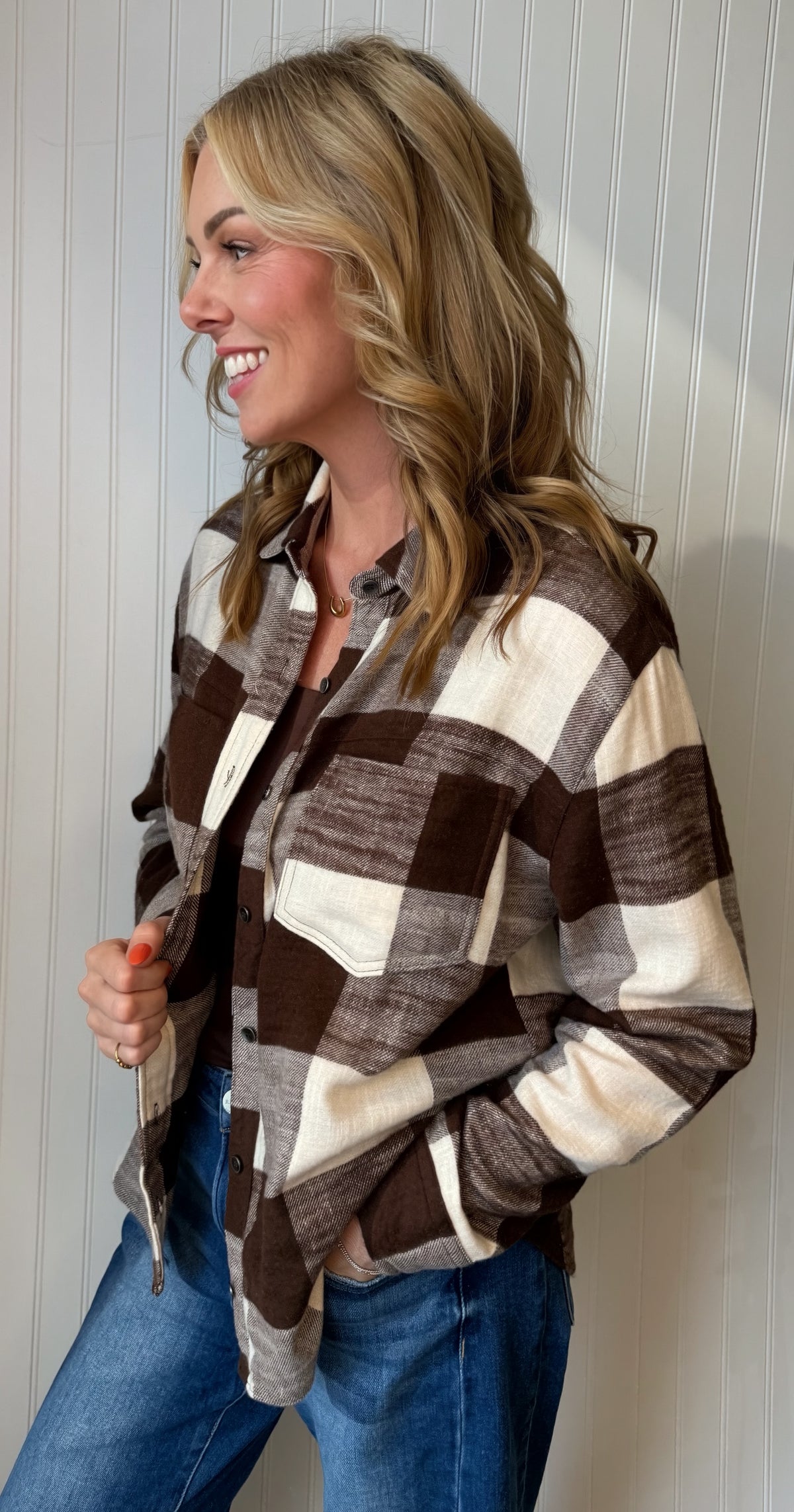A woman wearing a brown and cream buffalo plaid flannel shirt with blue jeans, standing against a white paneled wall.
