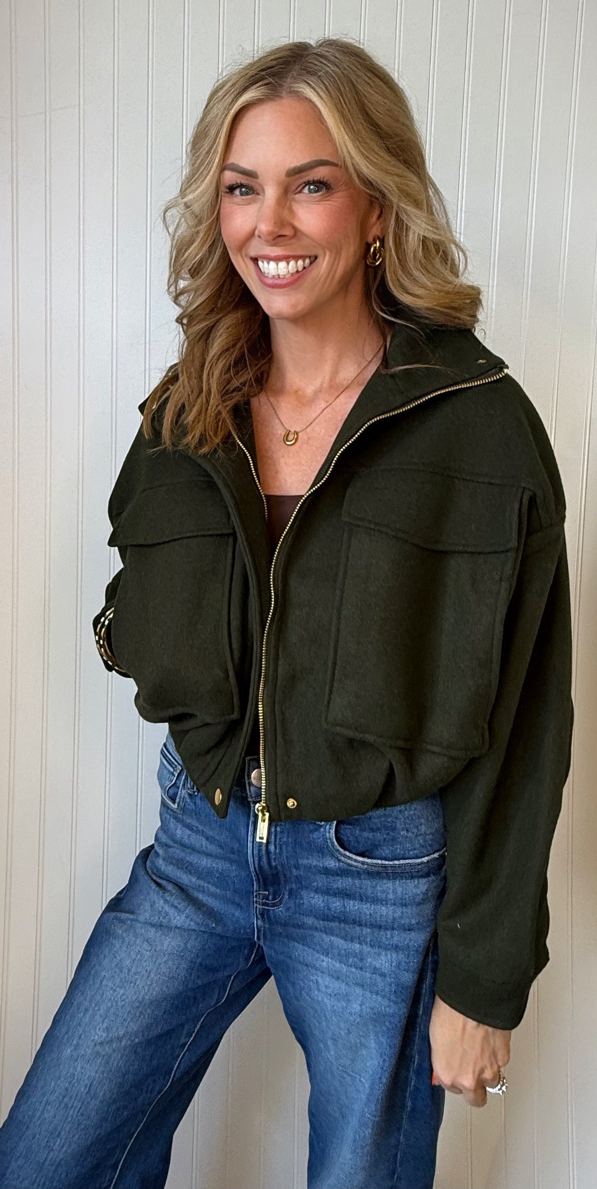 A woman wearing an olive green cropped zip-up jacket with front flap pockets and blue jeans, standing against a white paneled wall.