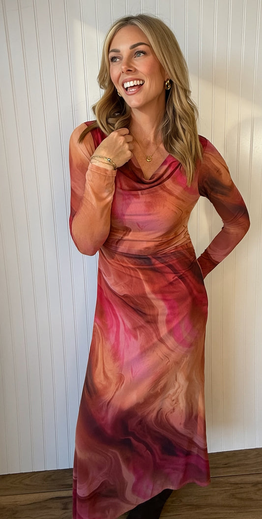 A woman modeling a long-sleeve cowl-neck dress with a pink, red, and orange marbled abstract print, standing against a white paneled wall.