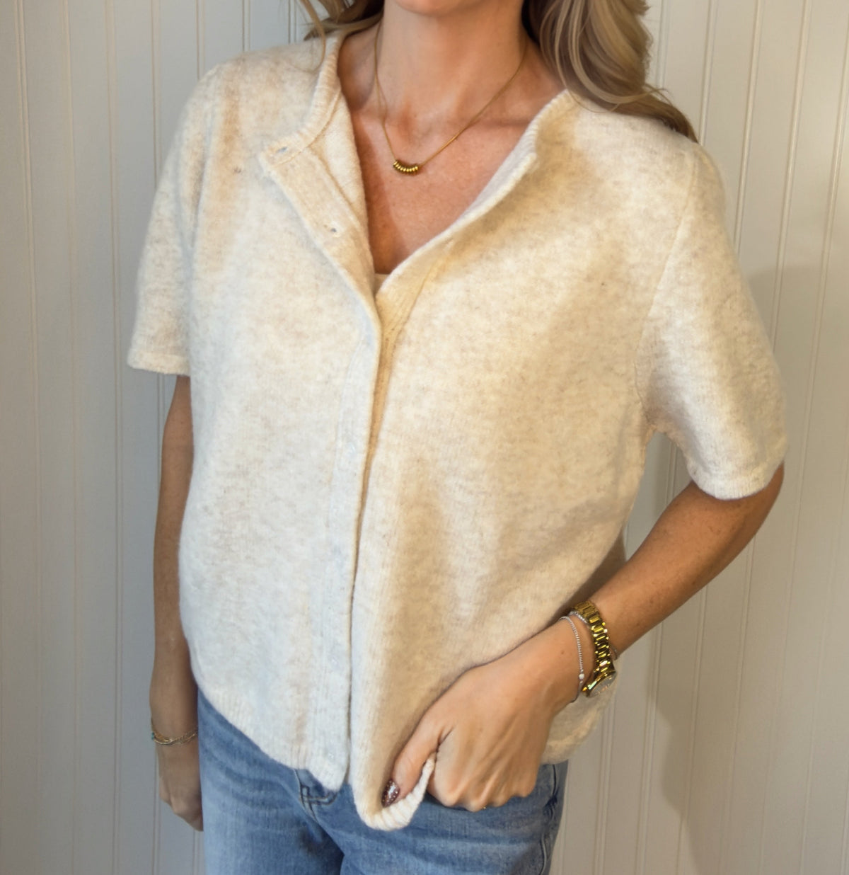 A smiling blonde woman wearing a short-sleeve cream-colored fuzzy knit top and blue jeans, standing against a white paneled wall.