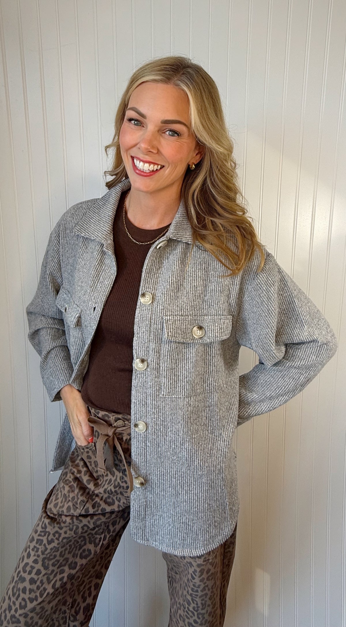 A smiling blonde woman wearing a light gray textured button-up overshirt over a dark brown top and leopard-print pants, standing against a white paneled wall.