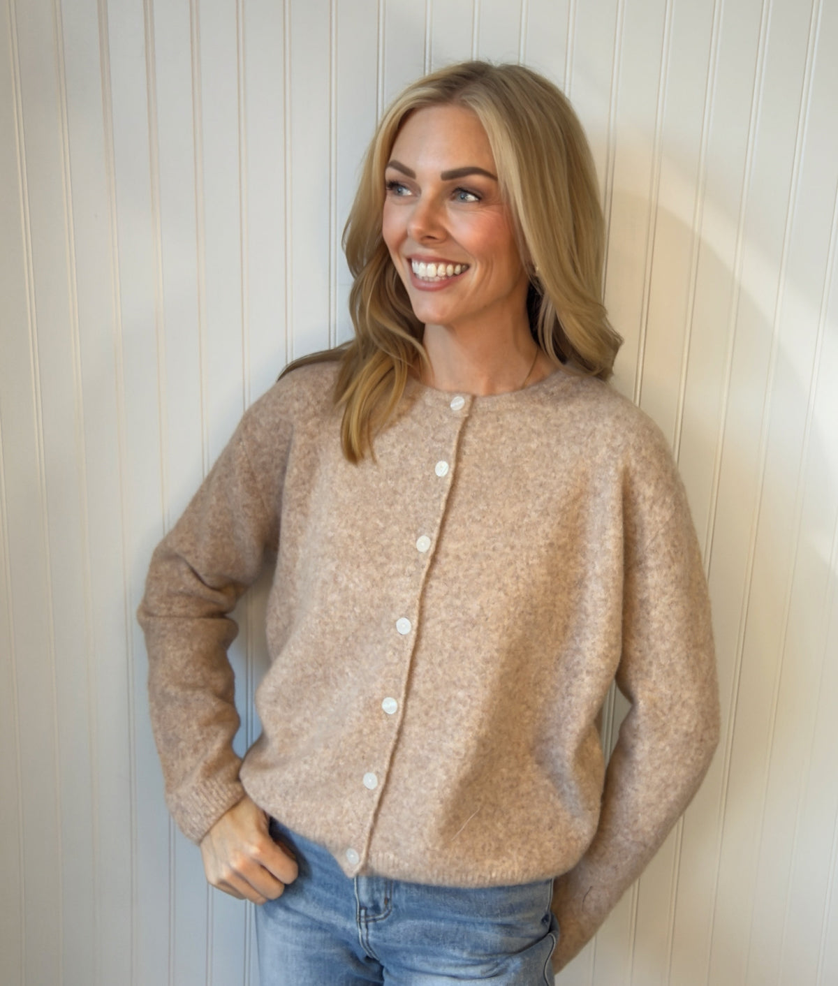A woman wearing a beige button-front knit cardigan and light blue jeans, standing against a white paneled wall.