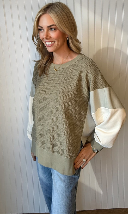 A woman modeling a textured knit sweater with a taupe body and contrasting cream sleeves, paired with light blue jeans, standing against a white paneled wall.
