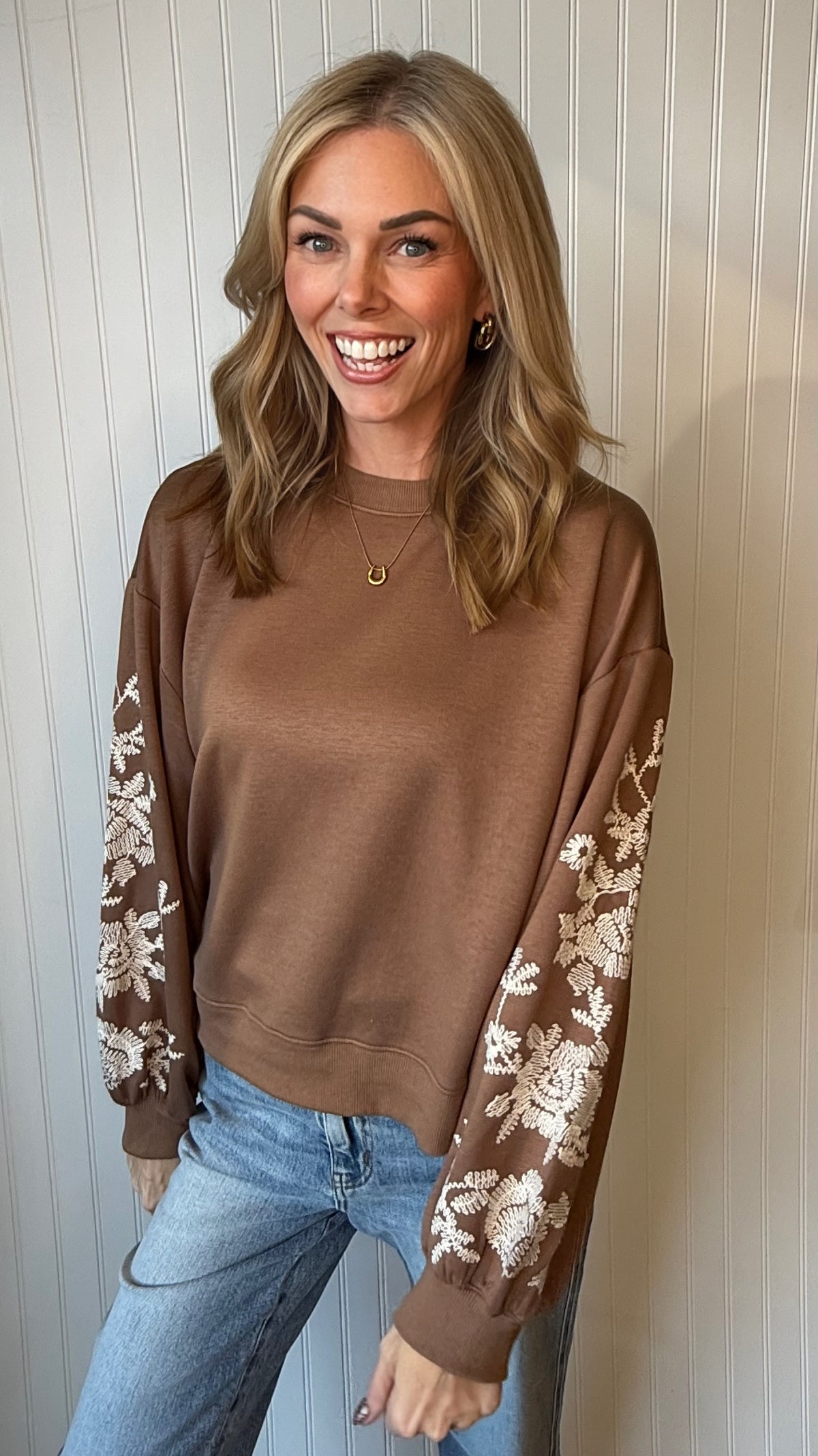 A woman smiling while wearing a brown crewneck sweatshirt with intricate white floral embroidery on the sleeves, paired with light blue jeans, standing against a white paneled wall.