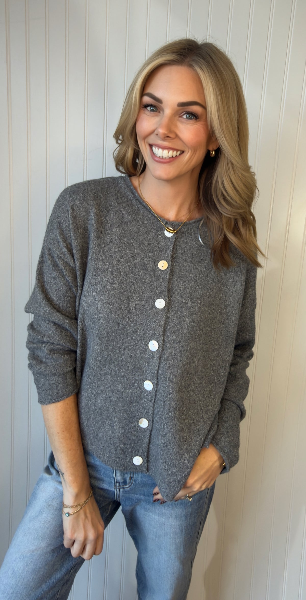 A smiling woman with blonde hair wearing a gray button-up cardigan and blue jeans, standing against a white paneled wall.