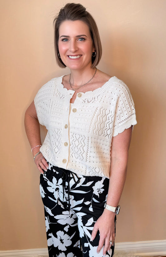 Woman wearing a white lace top with black floral pants against a beige wall.