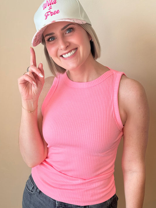 Woman wearing a pink tank top and white cap with 'Wild and Free' text, posing against a beige background.