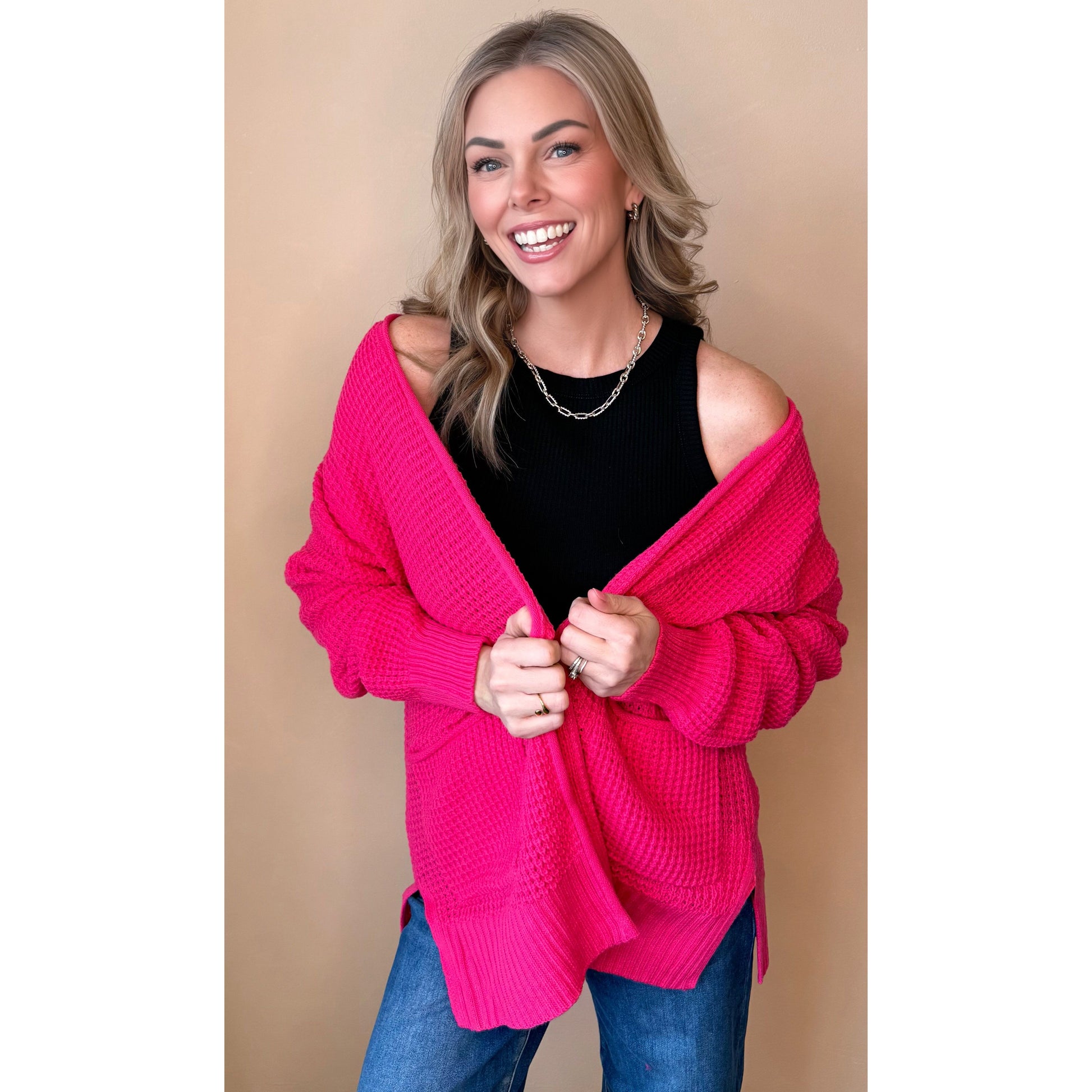 Woman wearing a bright pink cardigan over a black top and blue jeans on a beige background