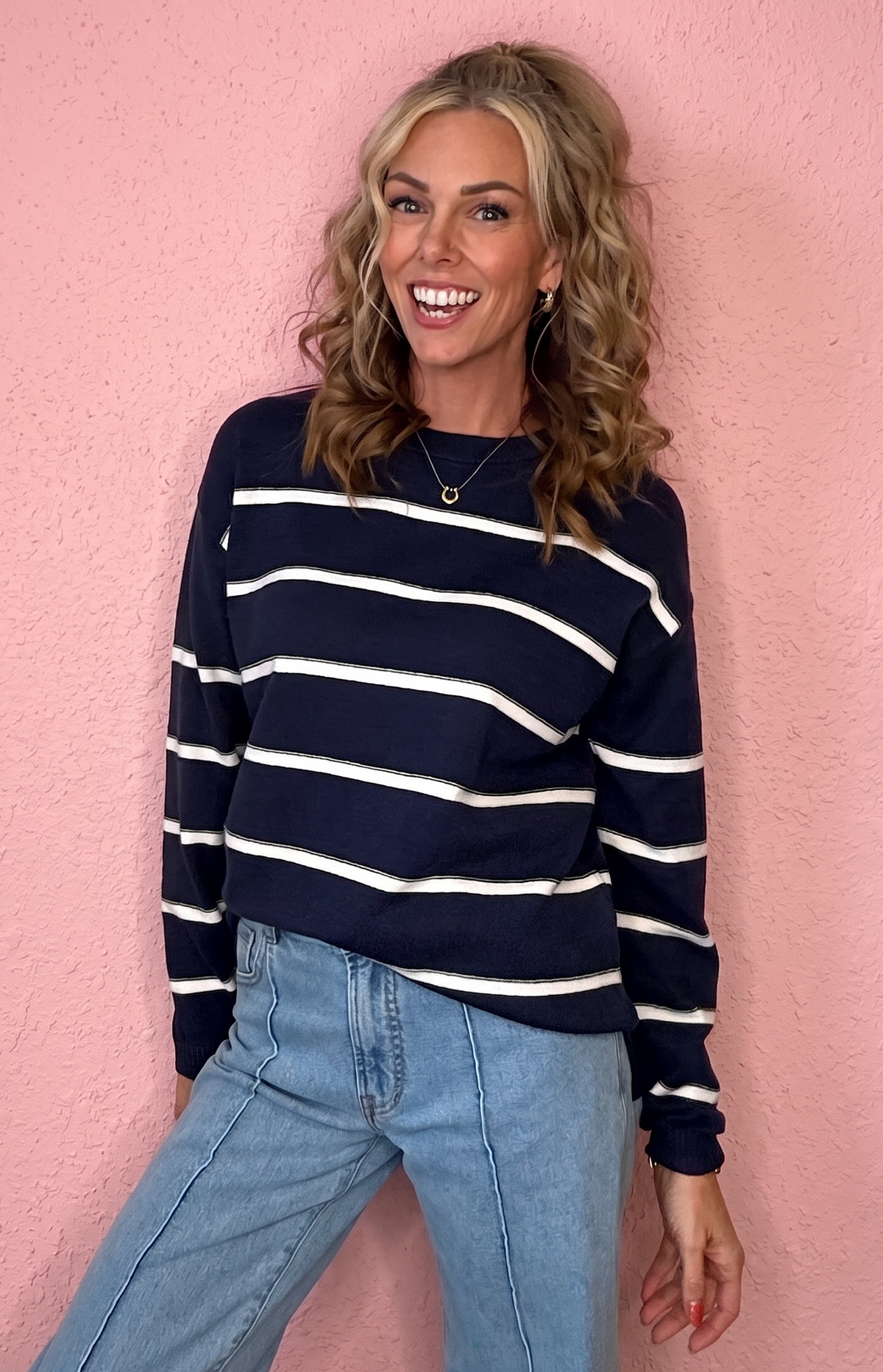 A woman smiling and posing in a navy blue and white horizontally striped long-sleeve knit sweater, paired with light-wash blue jeans, set against a textured pink wall.