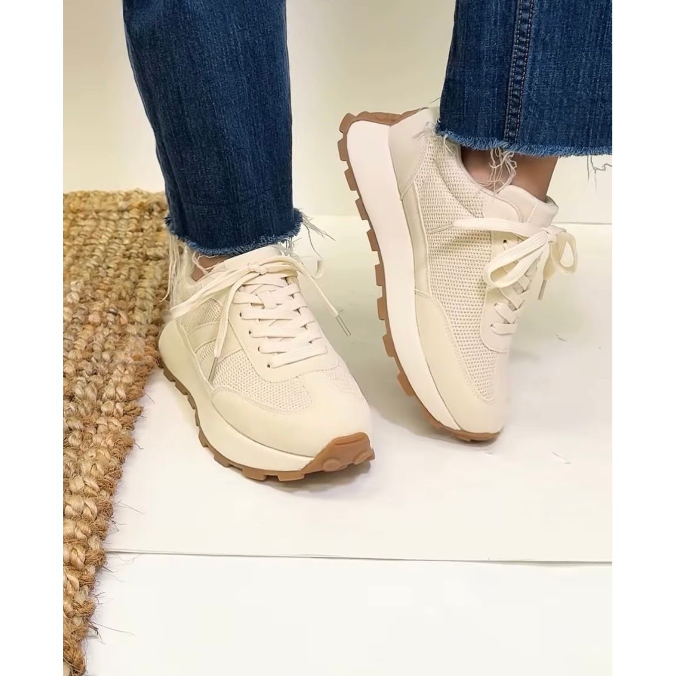 Beige sneakers with brown soles worn with blue jeans on a light wooden floor.