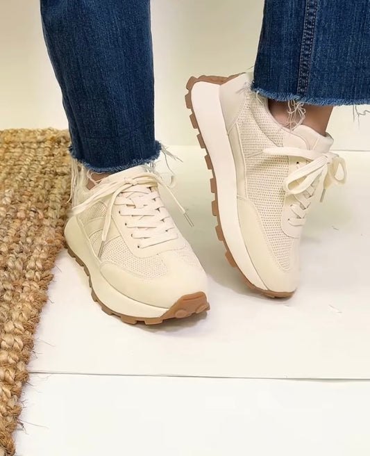 Beige sneakers with brown soles worn with blue jeans on a light wooden floor.