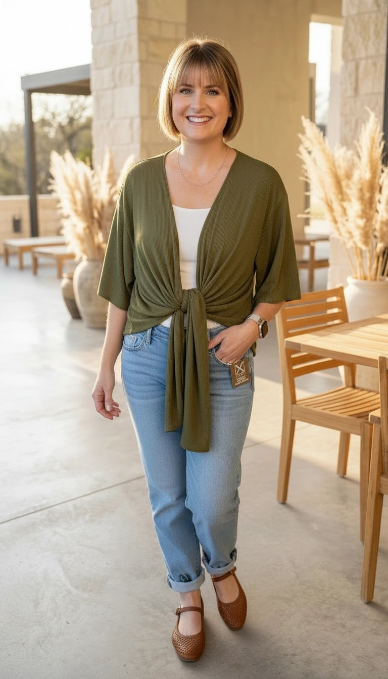 Woman wearing an olive green cardigan, white top, and blue jeans standing outdoors.