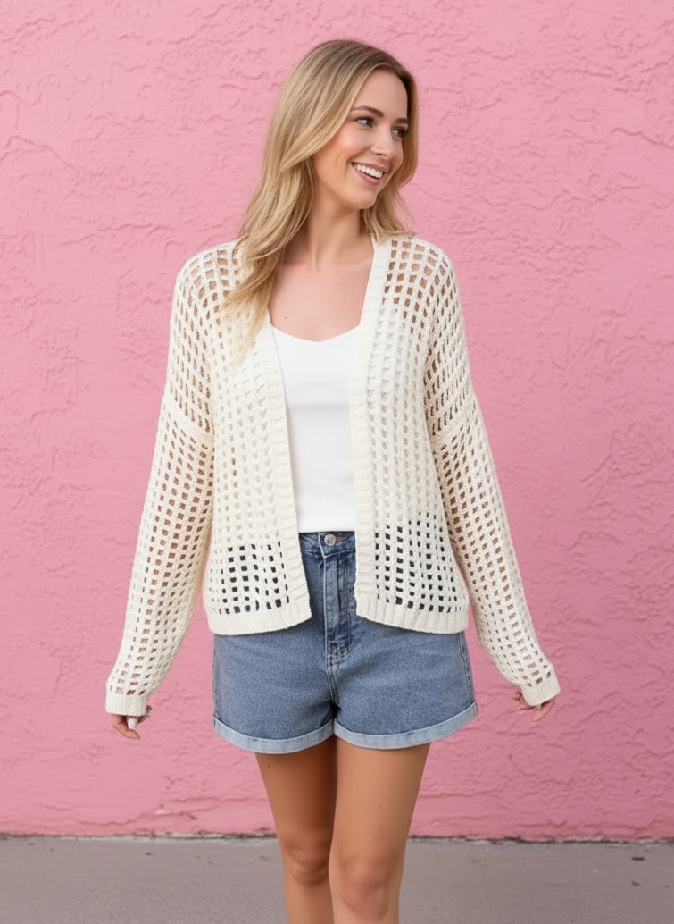 Woman wearing a white crochet cardigan over a white top and denim shorts against a pink wall.