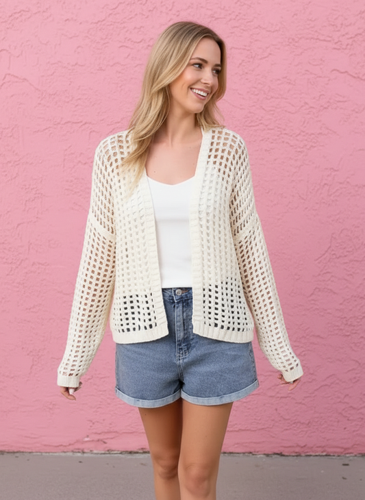 Woman wearing a white crochet cardigan over a white top and denim shorts against a pink wall.