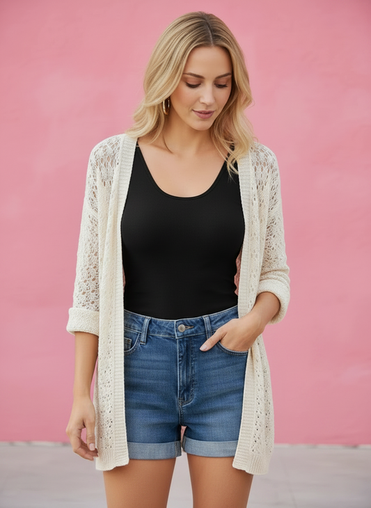Woman wearing a black top, denim shorts, and a beige cardigan against a pink background