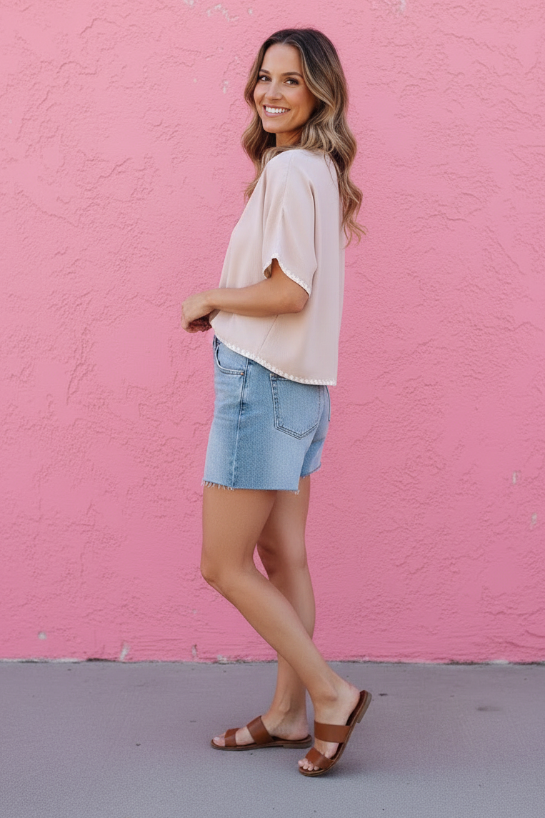 Woman in a light pink top and denim shorts standing against a pink wall.