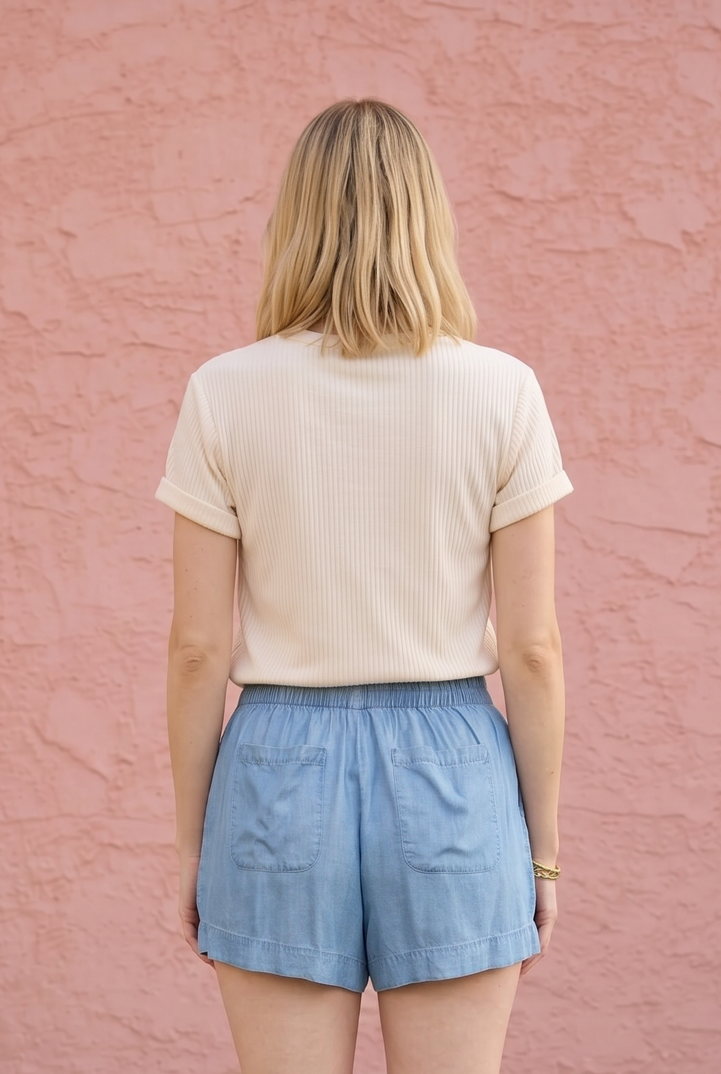 Person wearing a beige top and blue shorts standing against a pink wall.