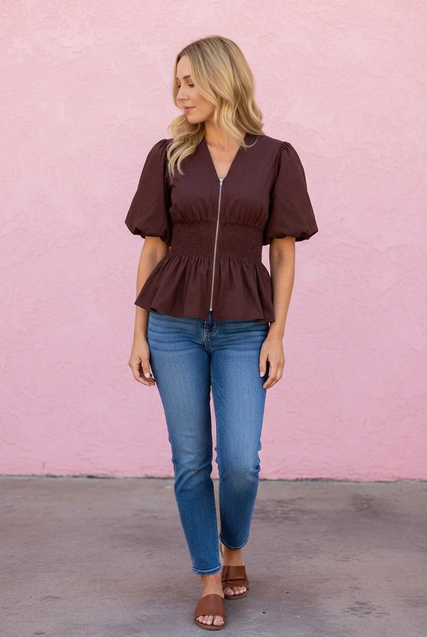Woman wearing a brown peplum top and blue jeans against a pink wall.