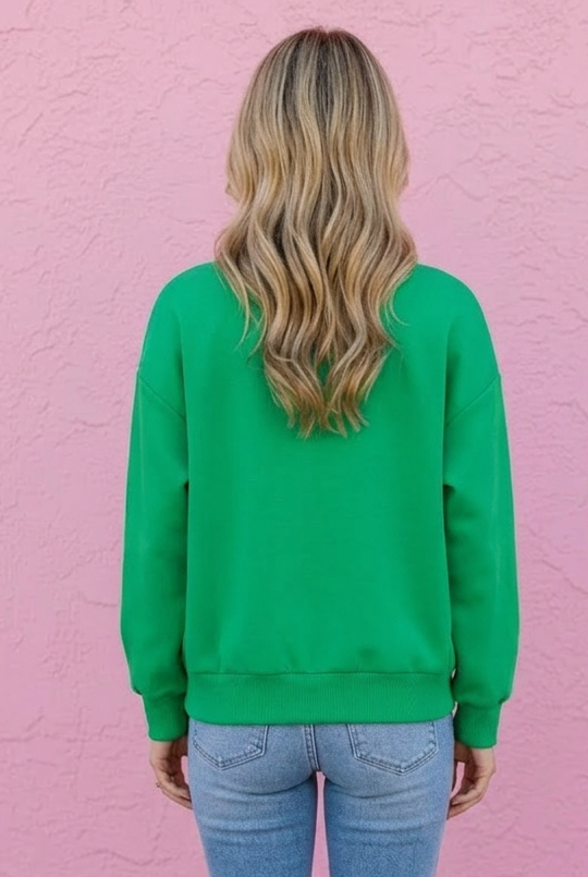 Person wearing a green sweatshirt and blue jeans standing against a pink wall.