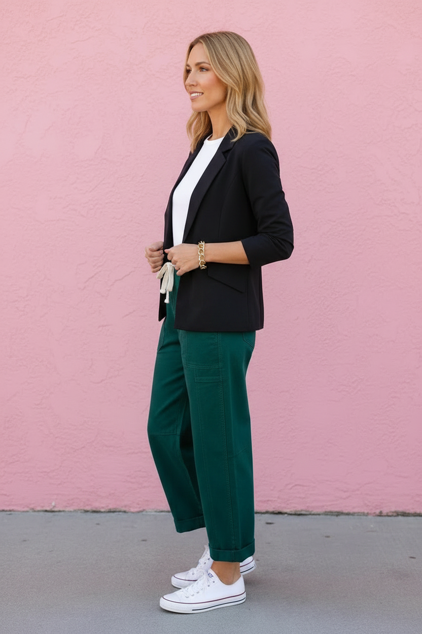Woman in a black blazer and green pants standing against a pink wall.
