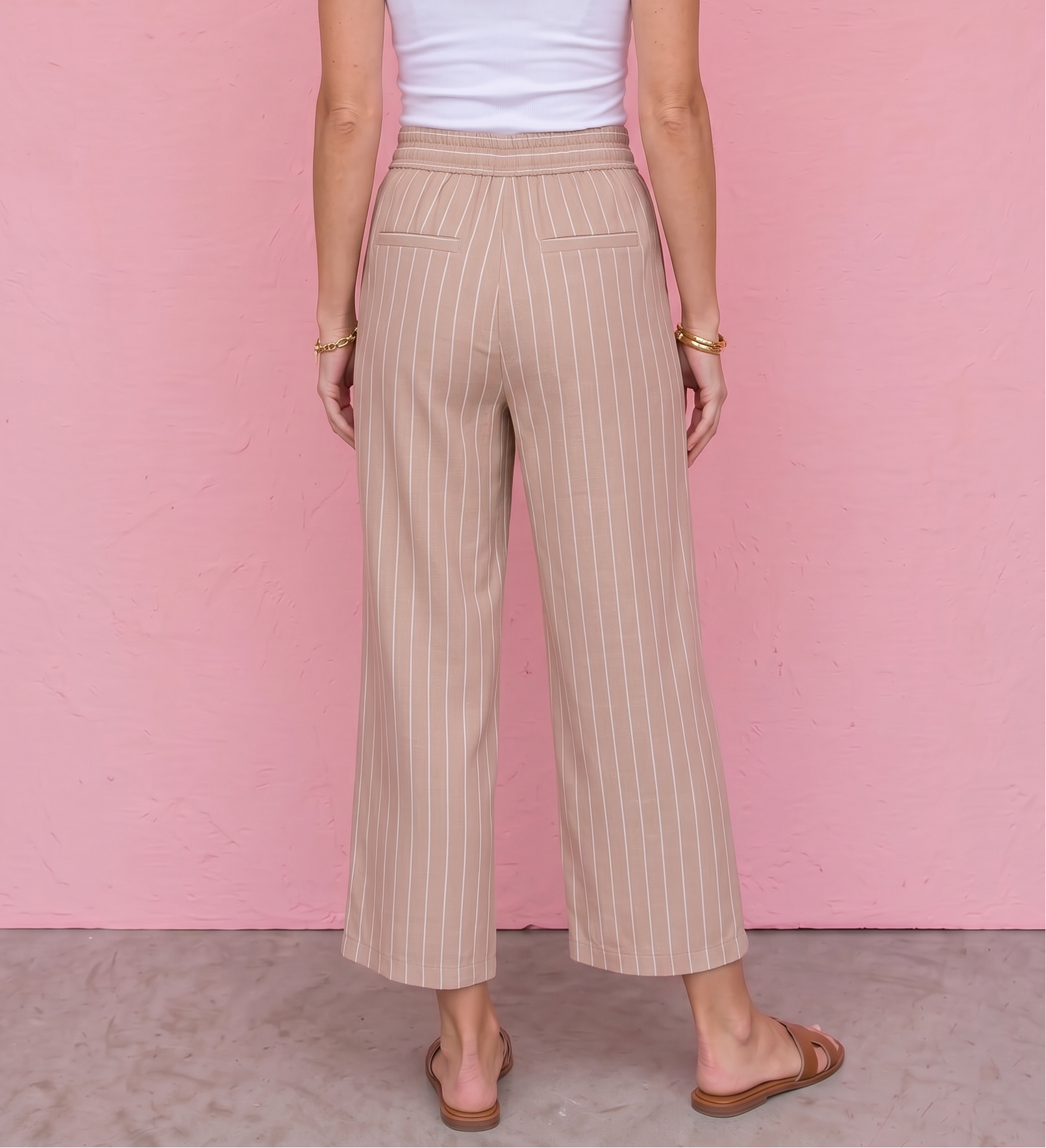 Woman wearing a white tank top and beige striped pants against a pink wall.