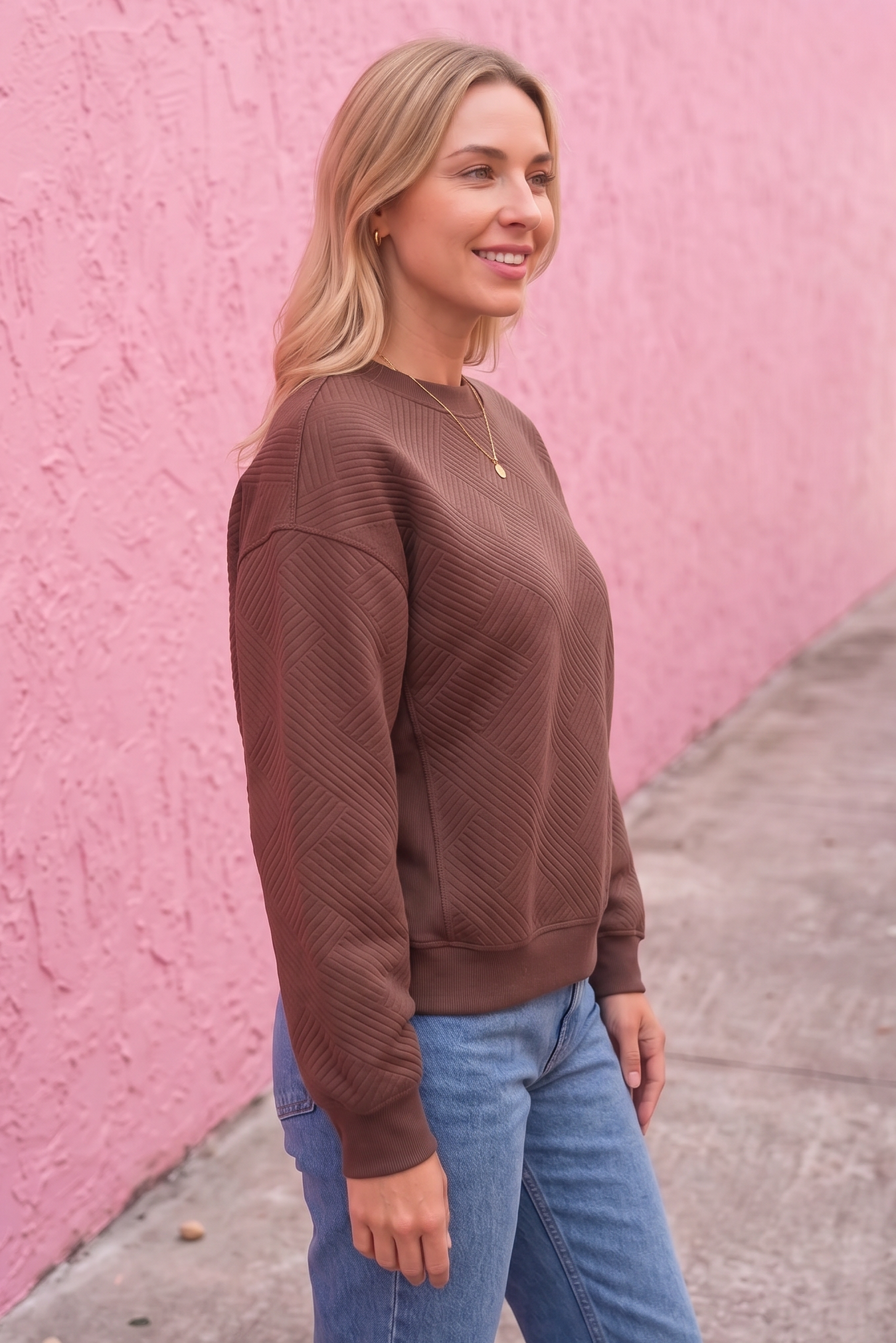 Woman wearing a brown sweater and blue jeans standing against a pink wall.