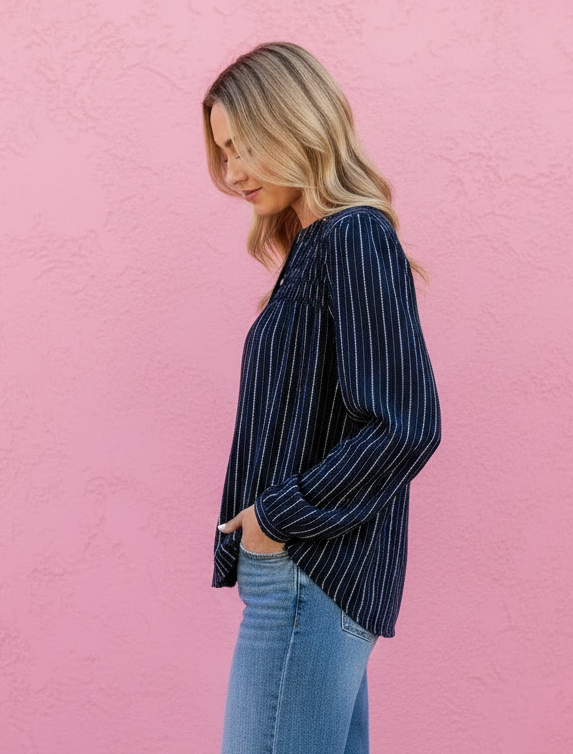 A woman stands in profile against a pink wall, wearing a navy blue long-sleeve top with thin white vertical stripes, wide-leg blue jeans, and brown sandals.