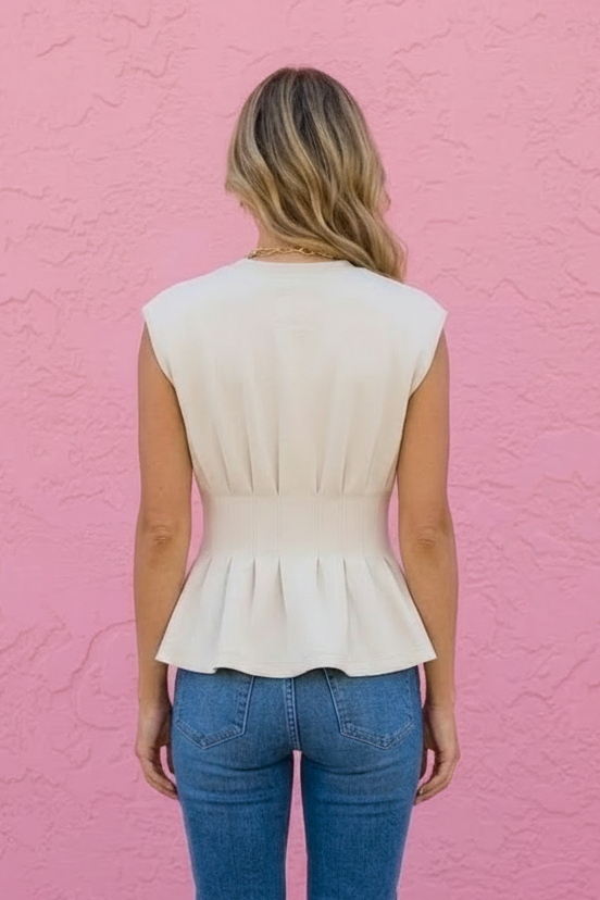 A woman viewed from behind wearing a cream-colored sleeveless peplum top and blue jeans, standing against a textured pink wall.