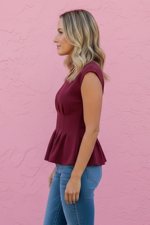 A woman stands in profile against a pink textured wall, wearing a sleeveless burgundy peplum top, blue jeans, and brown sandals.