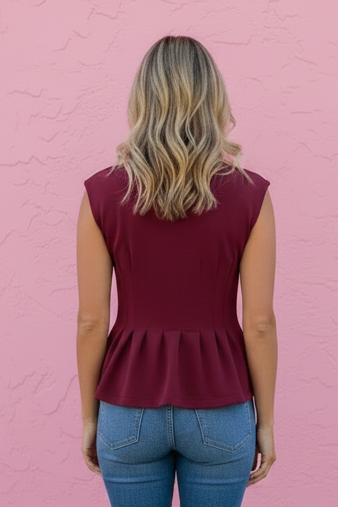 A woman viewed from behind wearing a sleeveless burgundy peplum top and blue jeans, standing against a textured pink wall.