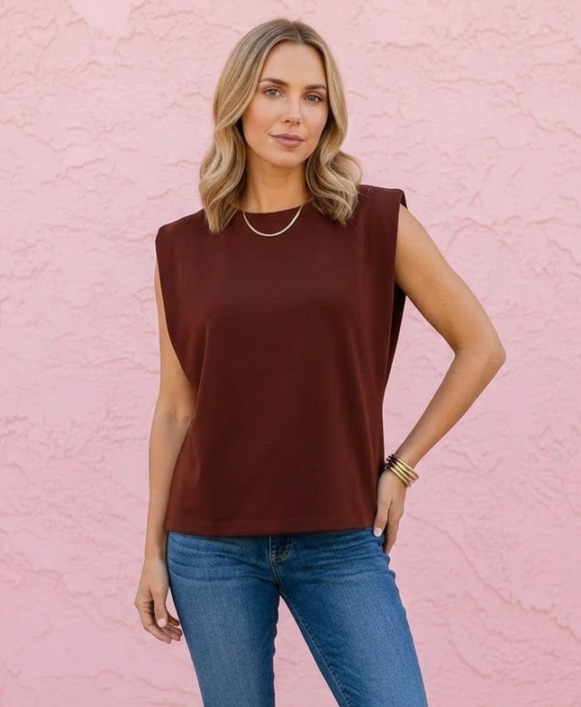 A woman modeling a sleeveless dark brown top with blue skinny jeans and brown sandals, standing against a textured pink wall.