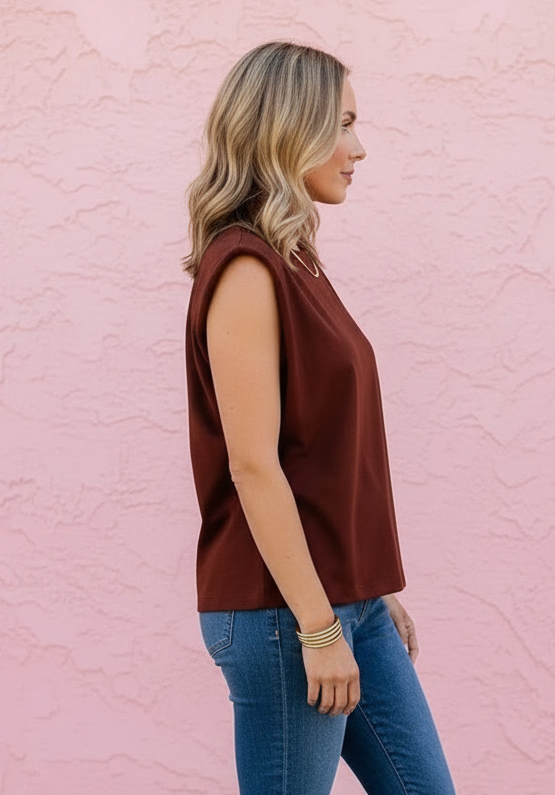 A woman in a sleeveless rust-brown top and blue skinny jeans stands in profile against a pink stucco wall, wearing brown slide sandals.