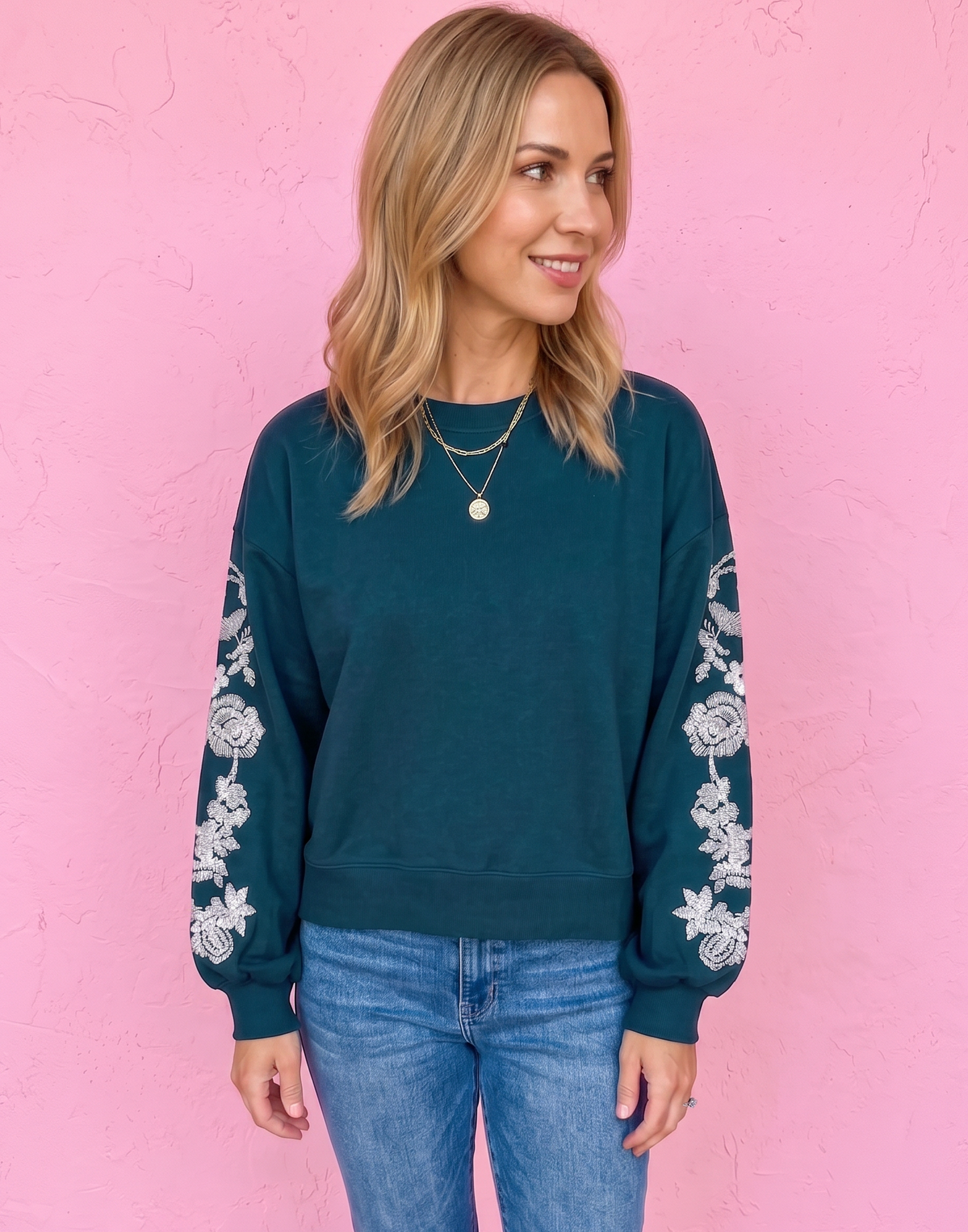 A woman wearing a dark teal crewneck sweatshirt with white floral embroidery on the sleeves, paired with blue jeans and brown sandals, standing against a pink wall.