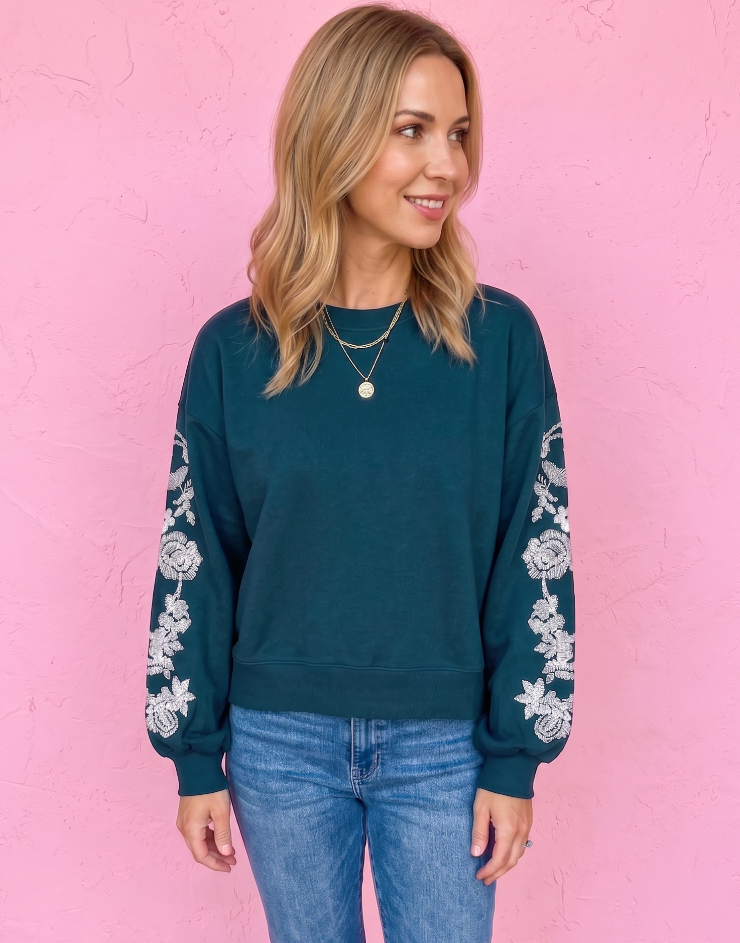 A woman wearing a dark teal crewneck sweatshirt with white floral embroidery on the sleeves, paired with blue jeans and brown sandals, standing against a pink wall.