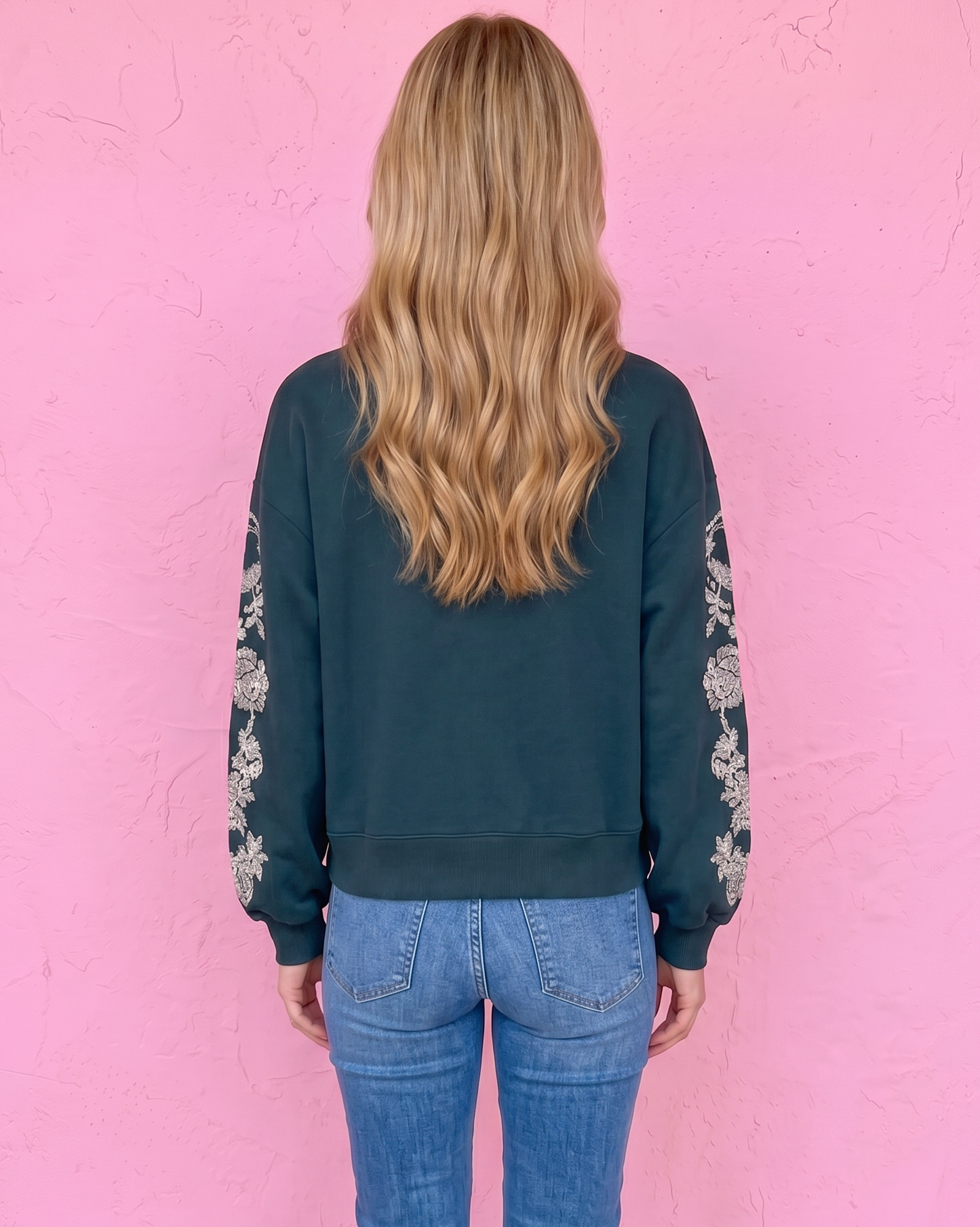 A woman viewed from behind wearing a dark teal green crewneck sweatshirt with white floral embroidery on the sleeves, paired with blue jeans and brown sandals, standing against a textured pink wall.
