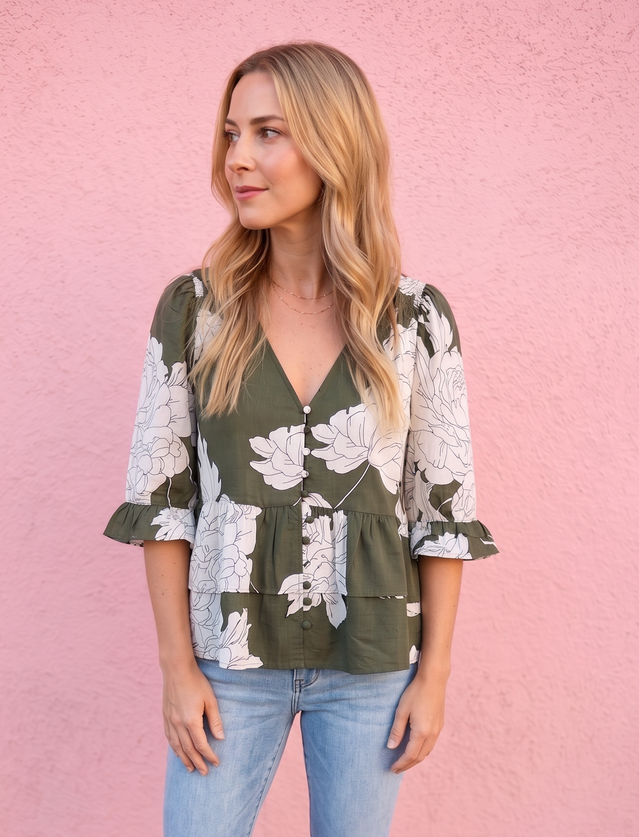 A woman stands against a pink wall wearing a green and white floral peplum blouse, light blue jeans, and brown boots.