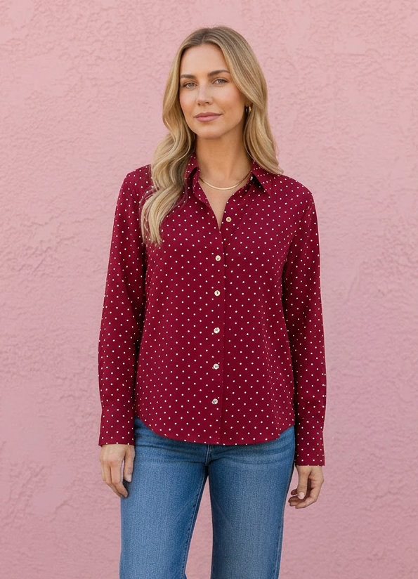 A woman stands against a pink stucco wall wearing a burgundy button-up shirt with a small white polka dot pattern, blue jeans, and black ankle boots.