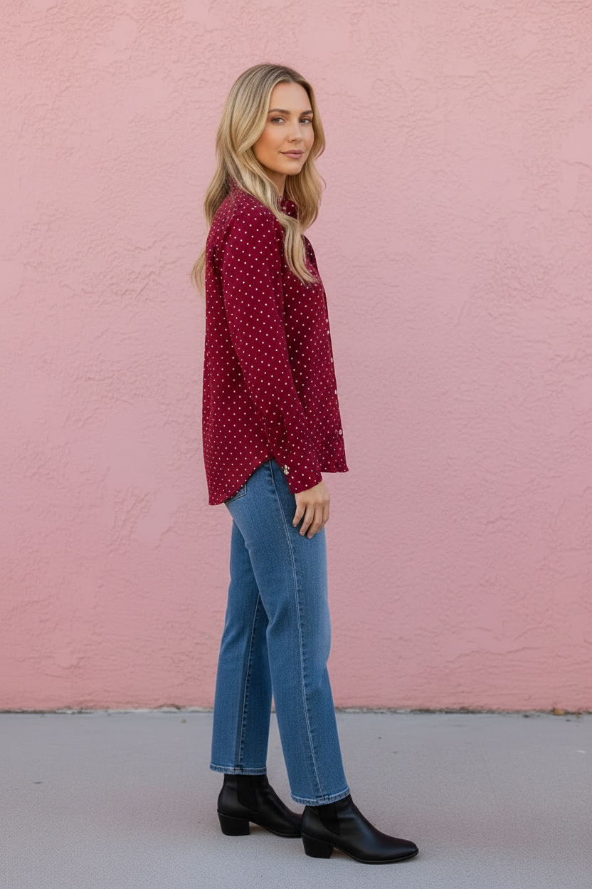A woman stands in profile against a pink stucco wall, wearing a burgundy long-sleeve blouse with a small white polka dot pattern, blue jeans, and black ankle boots.