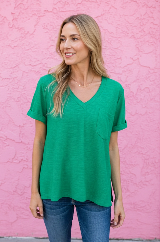 A woman wearing a bright green short-sleeve V-neck top and blue jeans stands in front of a textured pink wall.
