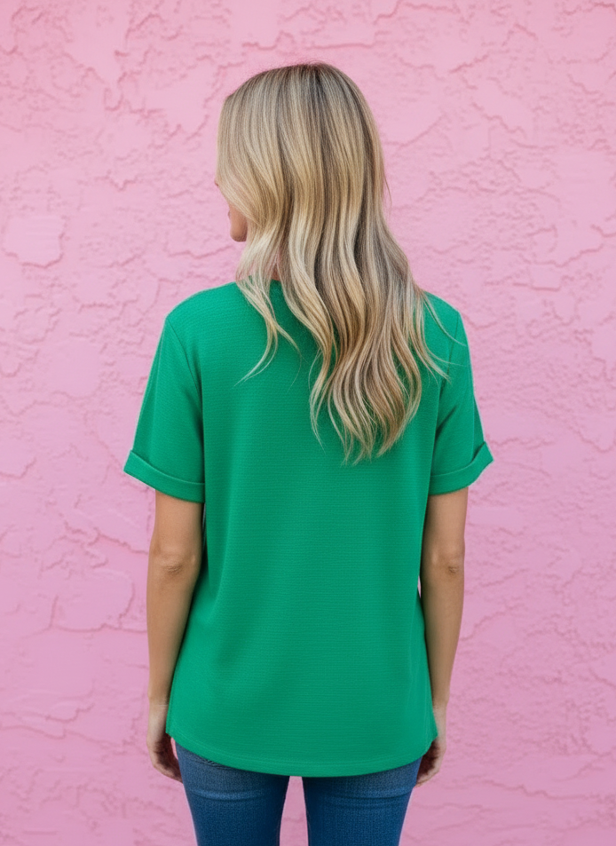 A woman with blonde hair is shown from behind wearing a bright green short-sleeve top and blue jeans, standing against a textured pink wall.