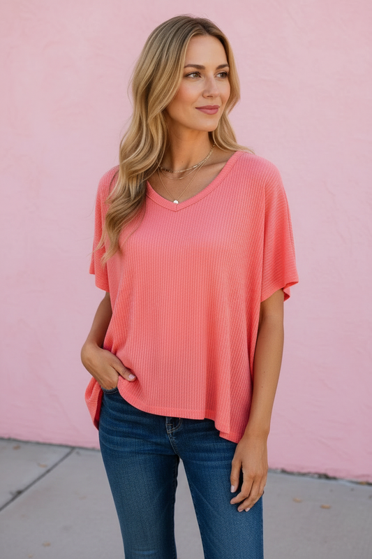 A woman wearing a coral pink ribbed short-sleeve V-neck top and blue jeans, standing against a pink wall.
