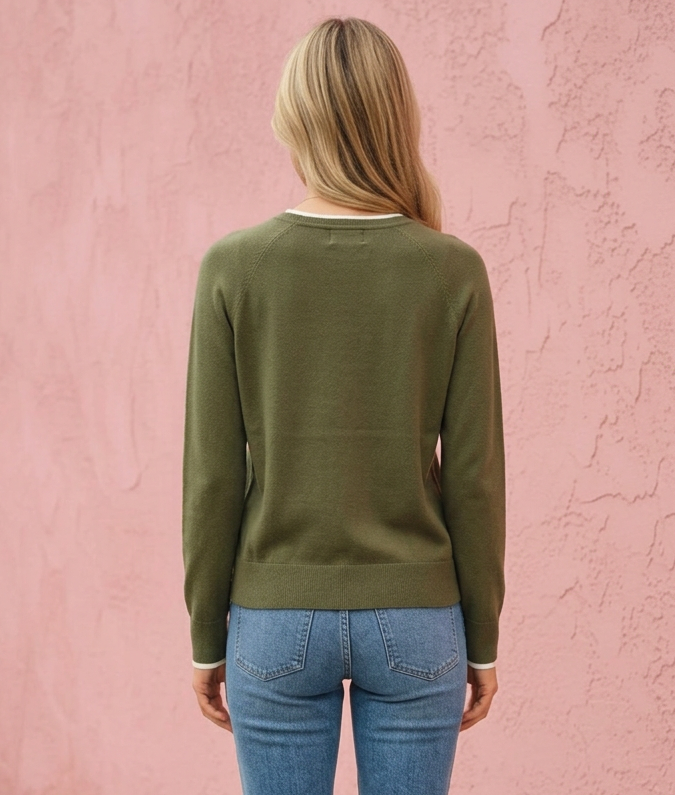 A woman viewed from behind wearing an olive green long-sleeve knit sweater, blue jeans, and black heels, standing against a textured pink wall.