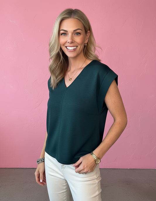 A smiling blonde woman wearing a dark teal green sleeveless V-neck top and white pants, standing against a textured pink wall.