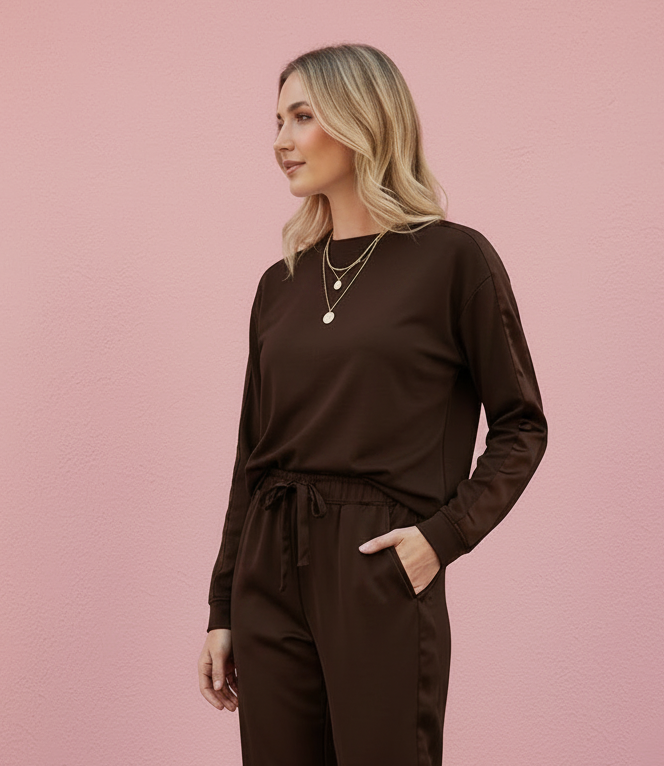 A woman modeling a dark brown long-sleeve top and matching drawstring jogger pants with side pockets, paired with white sneakers, set against a solid pink studio wall.