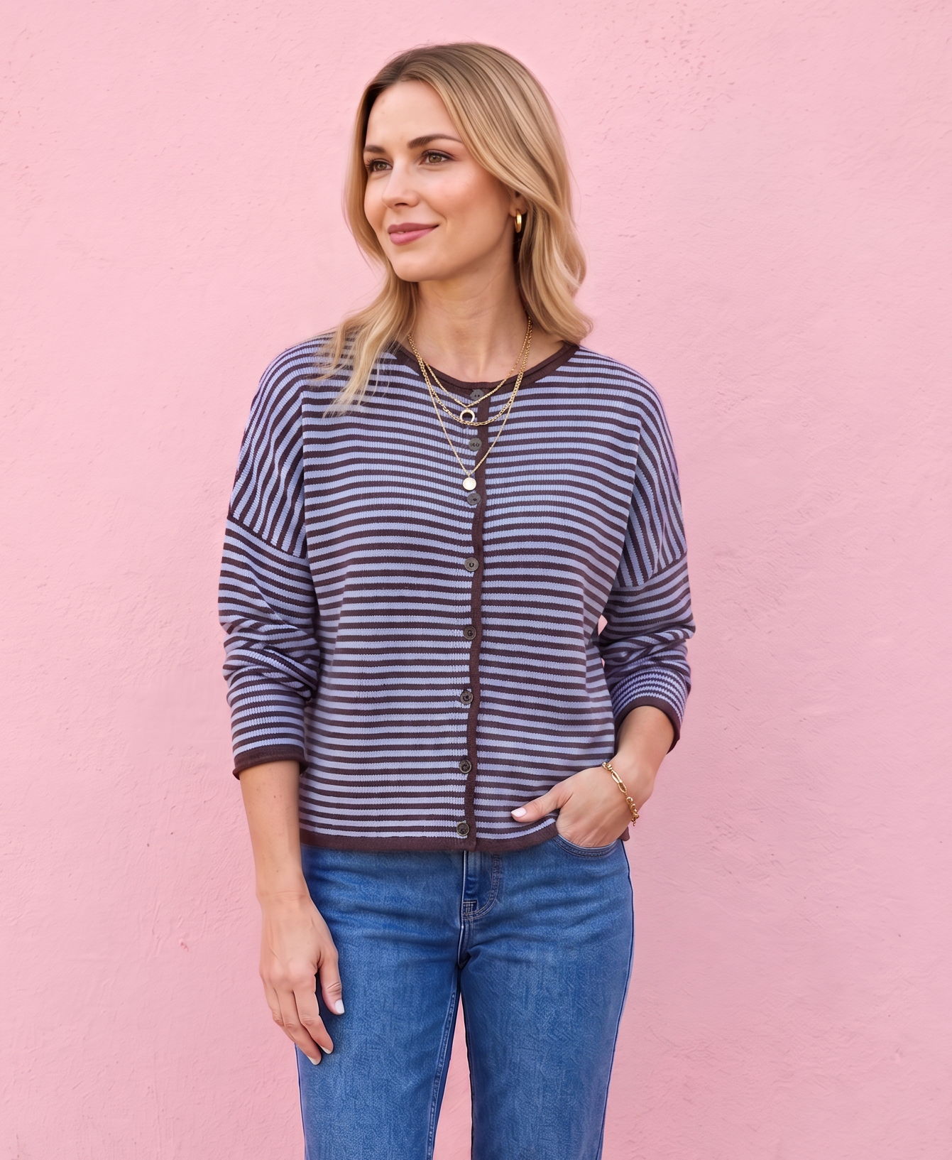 A woman stands against a pink wall wearing a navy and white striped zip-up cardigan, blue jeans, and dark brown boots.